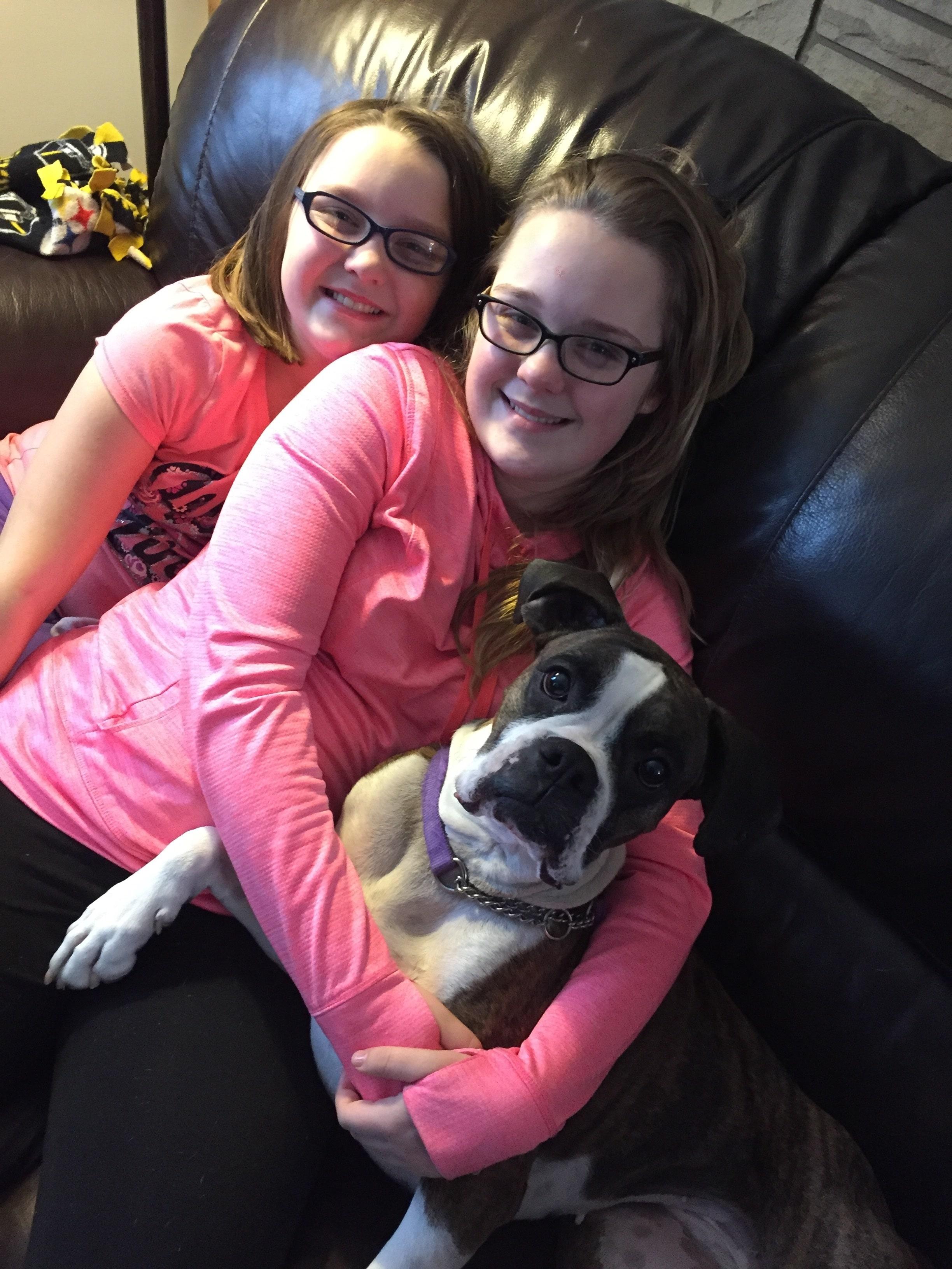 Two girls in pink on a couch sit hugging a black and white boxer.